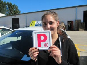 A successful local provisional student smiling from the window of the blue D-Tech vehicle while holding up her Red P-plate at the testing hub.