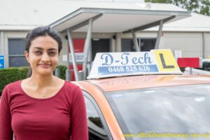 A successful young student giving a thumbs-up next to the D-Tech blue Corolla hybrid driving school training car.