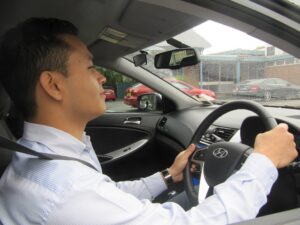 A successful young student giving a thumbs-up next to the D-Tech blue Corolla hybrid driving school training car.