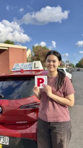 A happy female student holding up her new red P-plates next to a red D-Tech Driver Training car on a sunny day in Richmond.