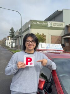 A smiling male student pointing at his new red P-plate while standing next to a red D-Tech driving school car in Richmond.