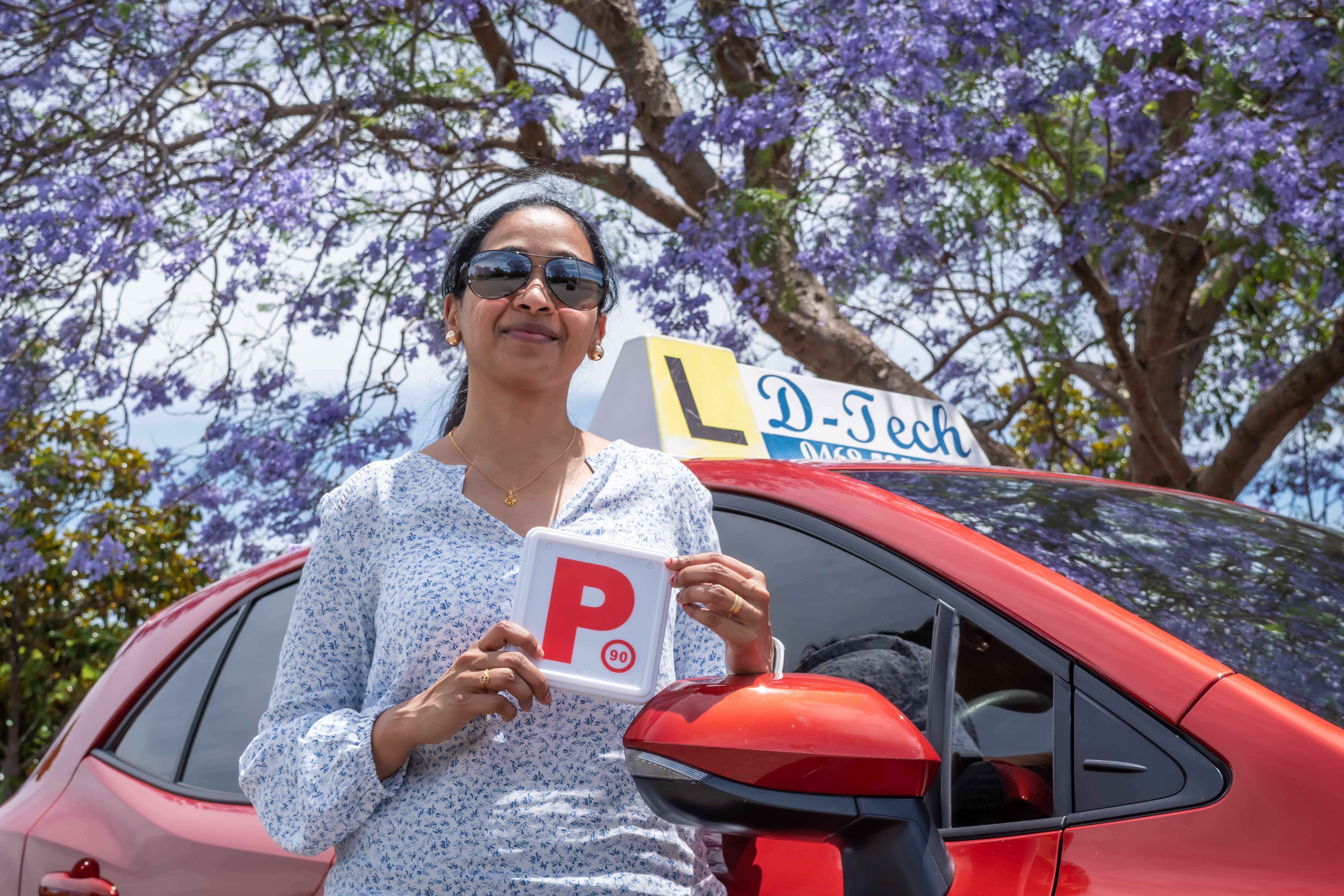 Happy student standing with P-plate under violet blooming trees next to red training car
