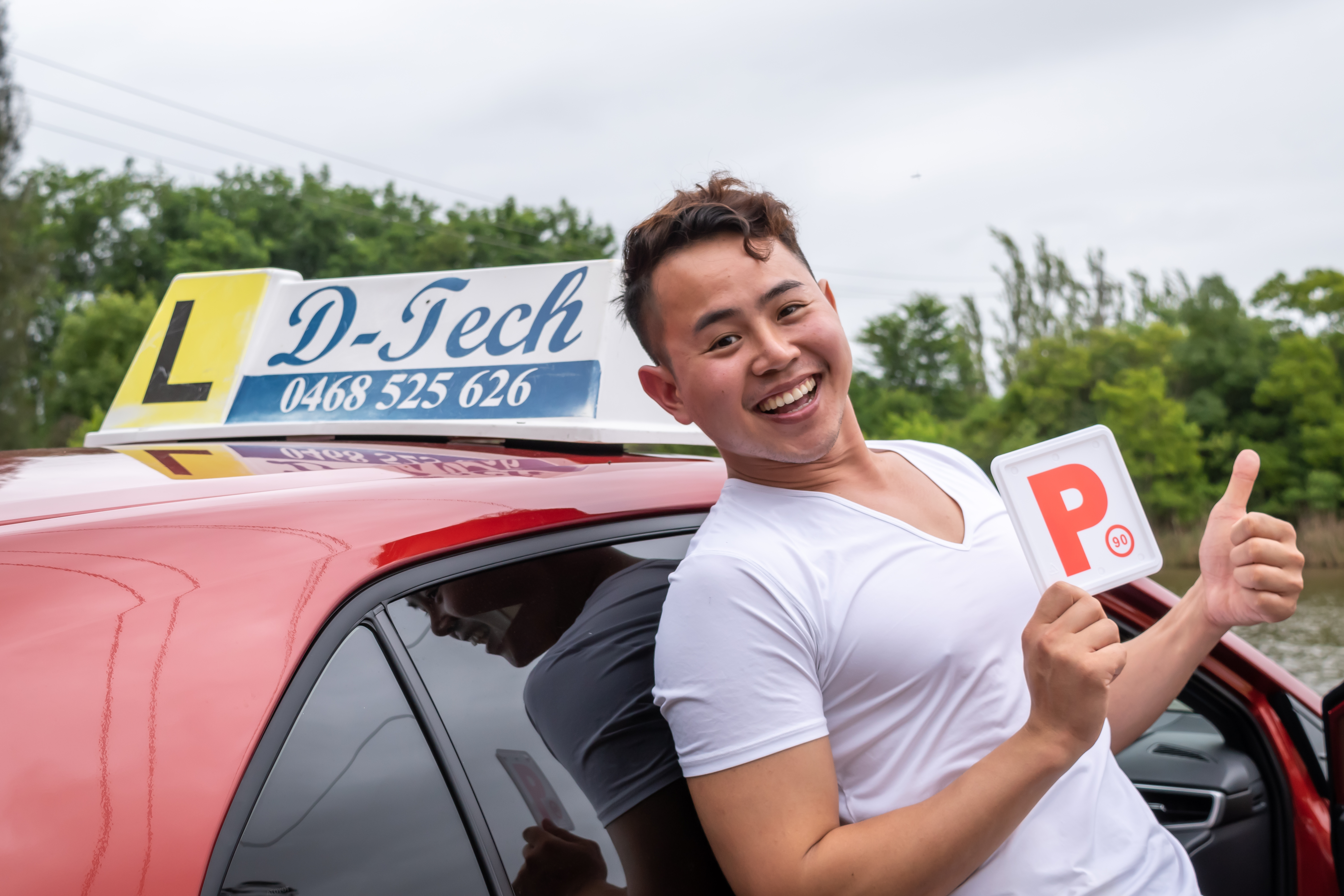 Male student leaning back on red car smiling holding fresh P-plate in celebration