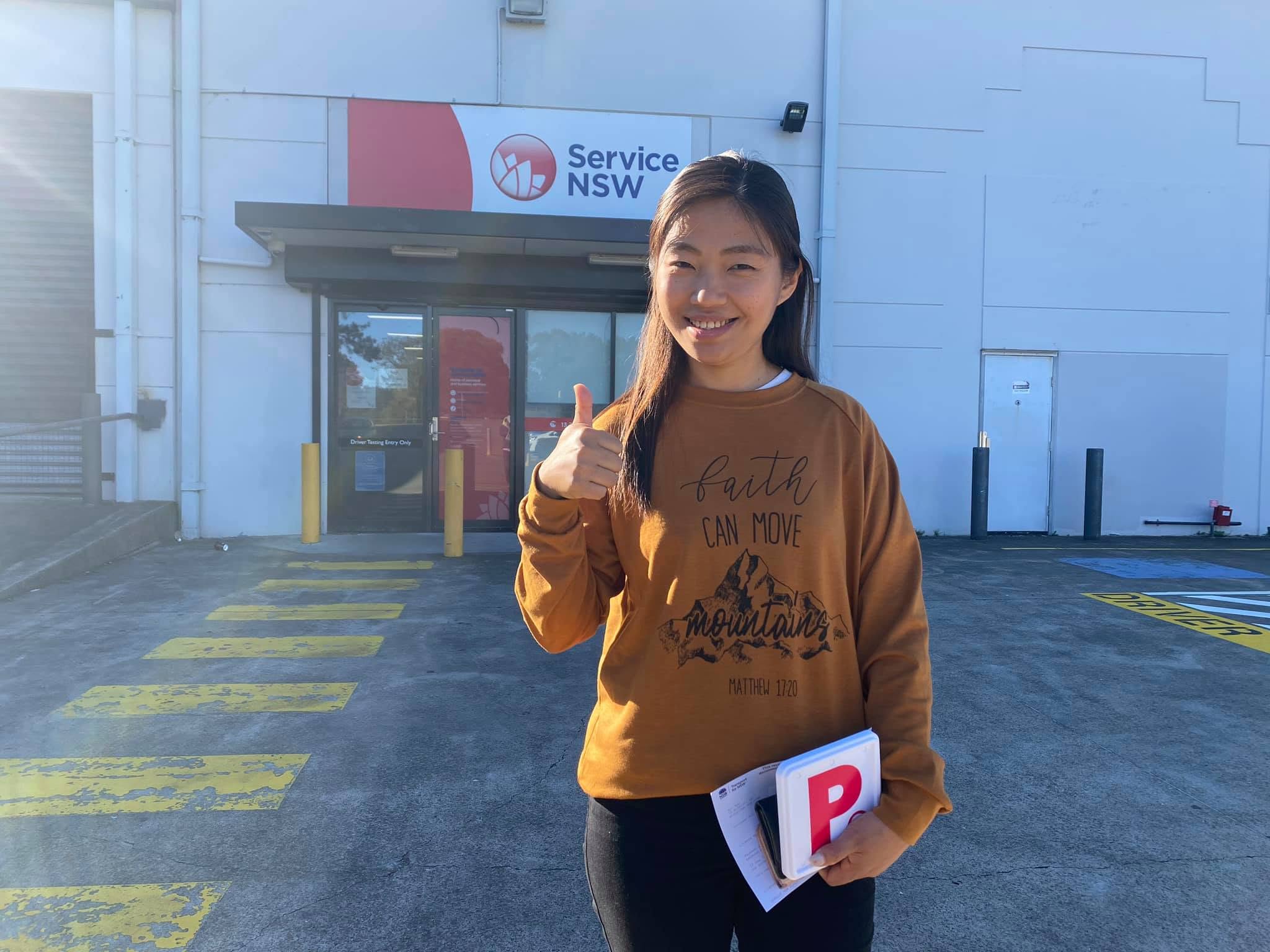 Happy student giving a thumbs up while holding her red P-plates after passing her practical driving test at the Service NSW Blacktown testing center.