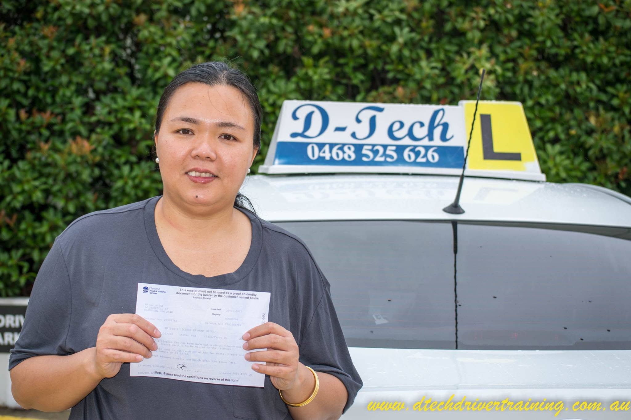 Successful student holding her official driving test pass receipt in front of a D-Tech Driver Training dual-control instruction vehicle.