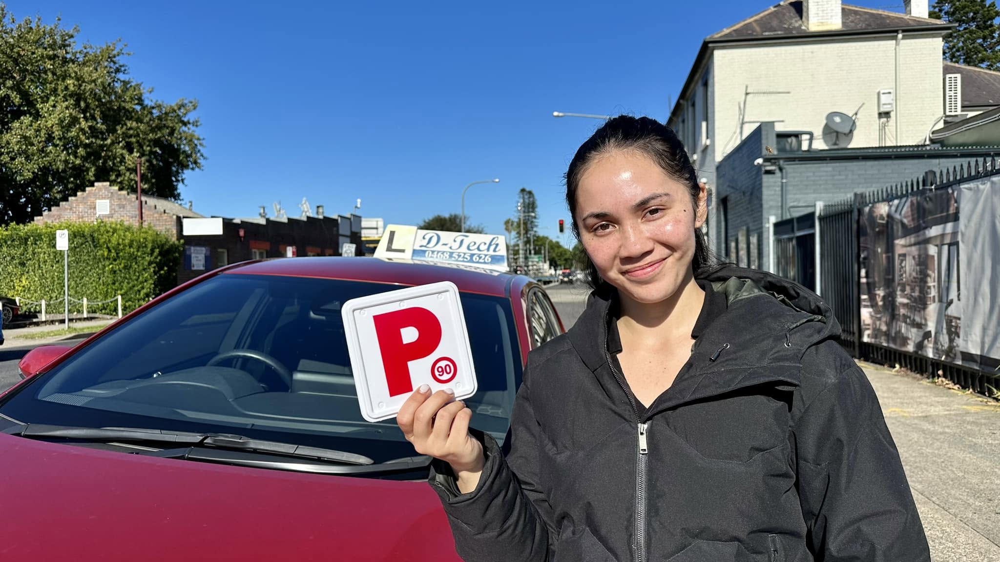 Smiling driving school student holding a red P-plate next to a D-Tech dual-control car after passing her practical driving test in Marsden Park.