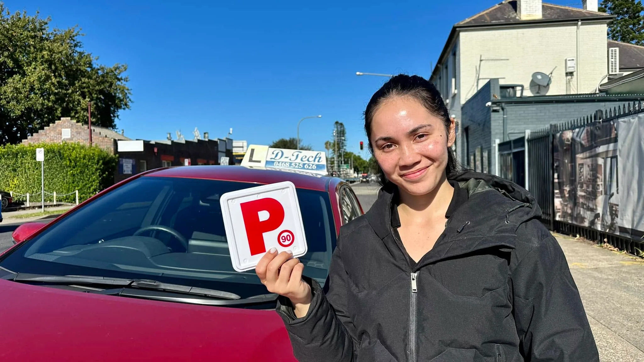 Smiling driving school student holding a red P-plate next to a D-Tech dual-control car after passing her practical driving test in Marsden Park.