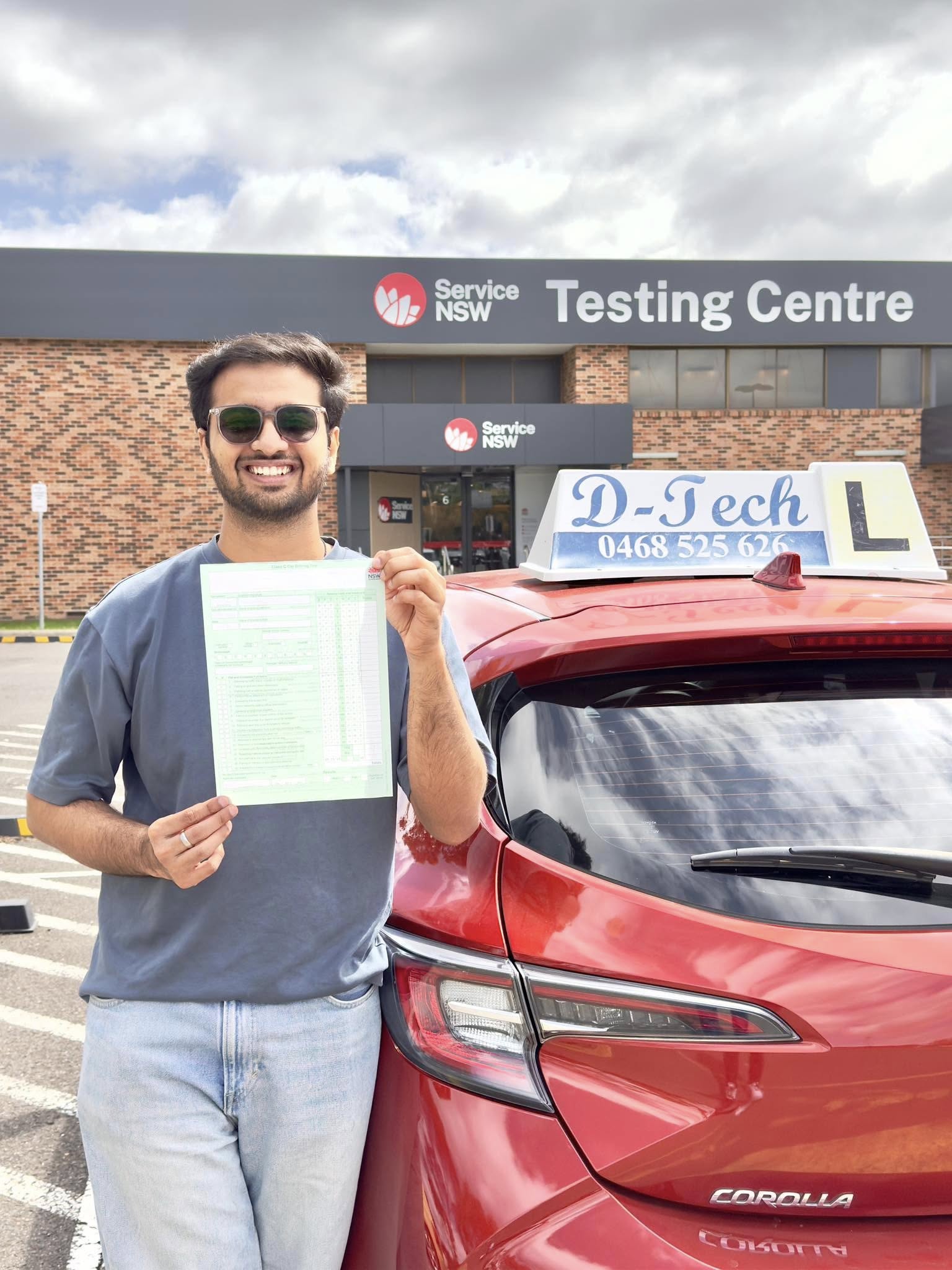 Smiling student holding a practical driving test pass sheet standing next to a D-Tech Driver Training car at the Service NSW St Marys testing centre with an expert driving instructor.