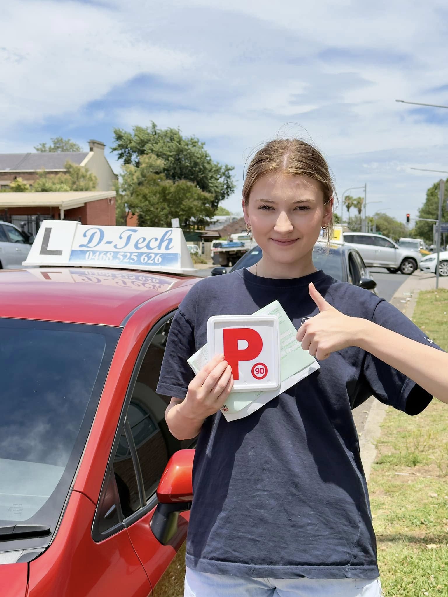 Successful student holding a red P-plate and giving a thumbs-up next to a D-Tech Driver Training car in South Windsor NSW.