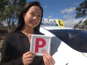 Smiling young blonde woman celebrating passing her driving test and holding red P-plates next to a white D-Tech driving school car.