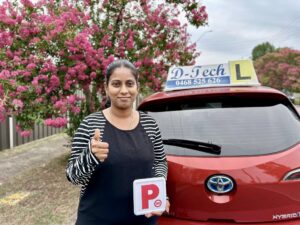A smiling female student standing next to a red D-Tech driving school car holding up her new provisional red P-plate in Tallawong.