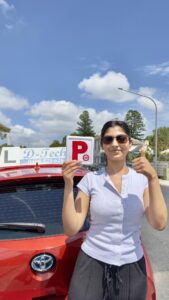 A happy female student holding up her new red P-plates next to a red D-Tech Driver Training car on a sunny day in Richmond Service NSW.