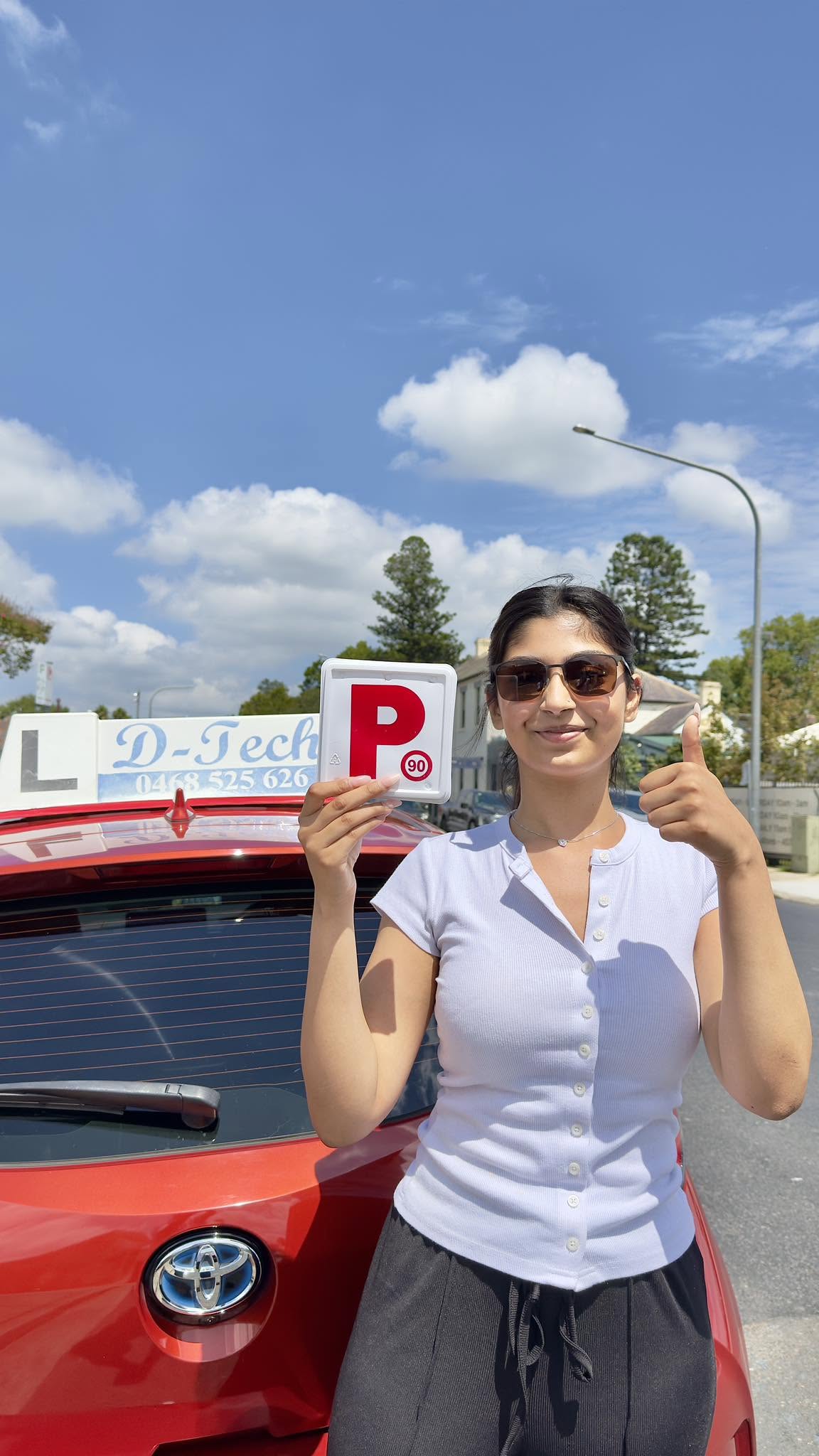 A happy female student holding up her new red P-plates next to a red D-Tech Driver Training car on a sunny day in Richmond Service NSW.