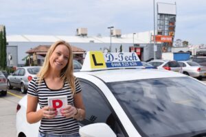 Smiling young Asian woman celebrating her practical driving test success with a red P-plate by a D-Tech car.