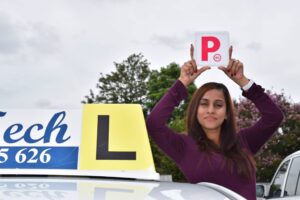 Successful male learner driver smiling and holding up a red P-plate next to a white D-Tech instructor vehicle.