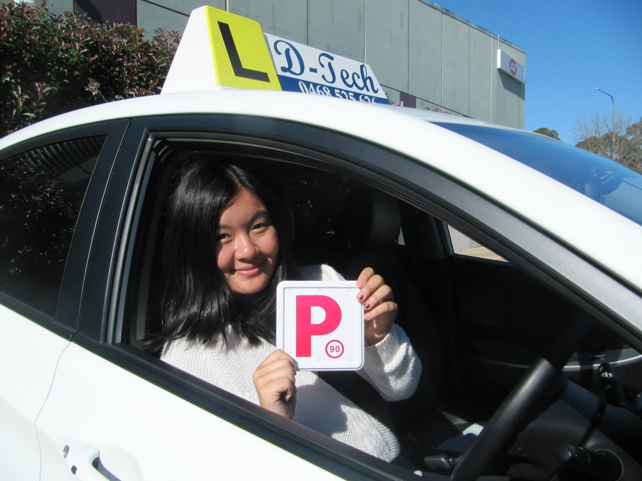 Student smiling behind the wheel inside a D-Tech Driver Training car holding her red P-plate after passing her test in Blacktown.