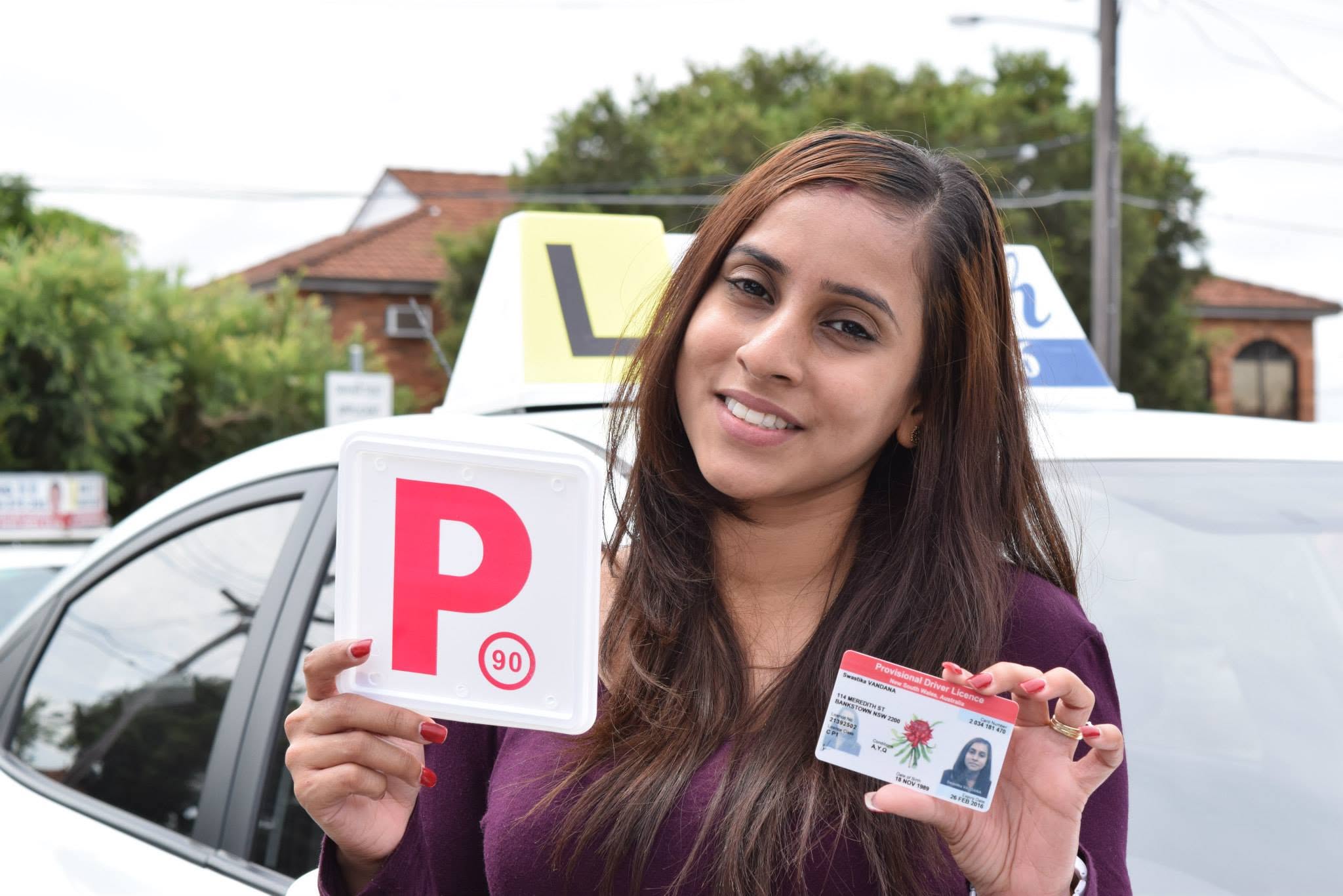 Happy student giving a thumbs up while holding her red P-plate in front of a D-Tech Driver Training dual-control car.