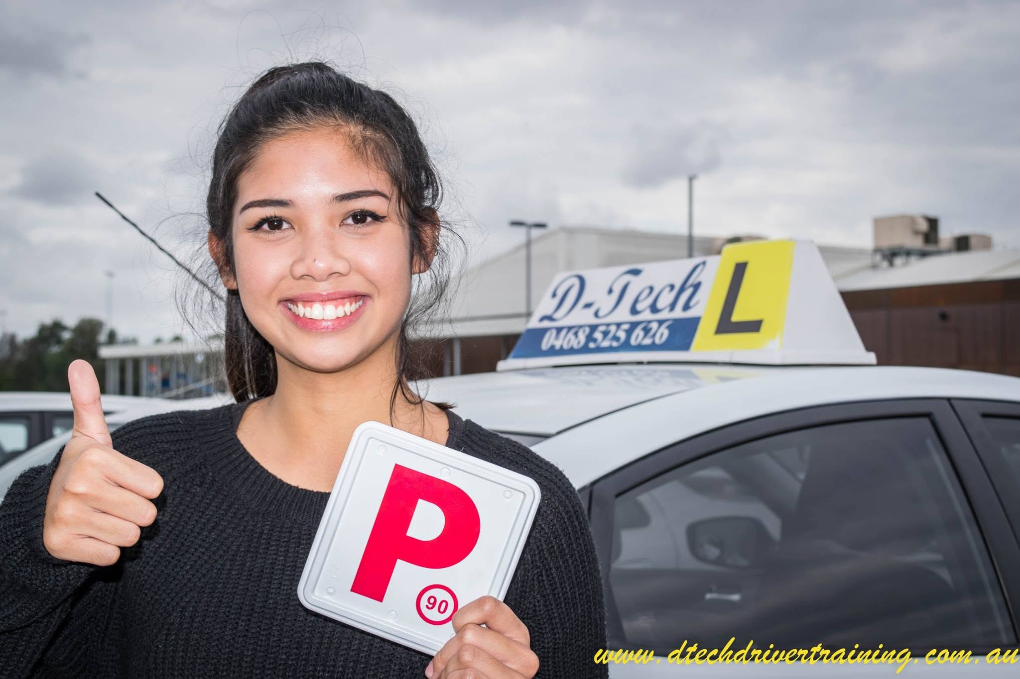 Student Monica giving a thumbs up while holding her red P-plate outside D-Tech Driver Training car after passing her driving test.